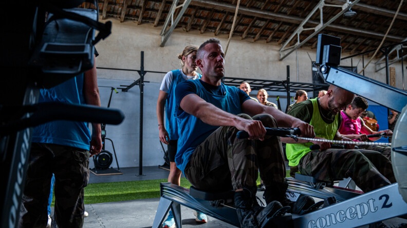 Trois hommes se défient au rameur dans une salle de sport, entourés par des personnes en treillis ou tenue de sport