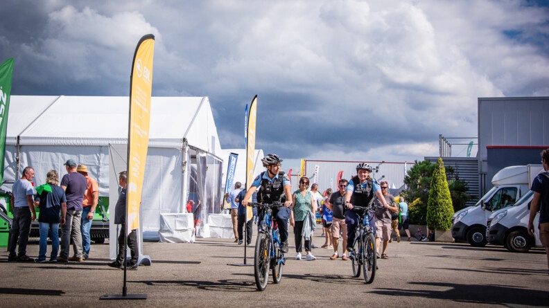 Deux gendarmes à vélo en patrouille autour des abords d'un salon. De part et d'autres des visiteurs, quelques camions et tentes d'exposition.