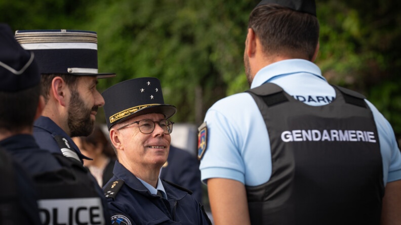 Le Major G&eacute;n&eacute;ral de la Gendarmerie Nationale &eacute;change, souriant,  avec un gendarme, que l'on voit de dos. Ce dernier porte un polo bleu ciel et un gilet pare balles noir rev&ecirc;tant l'inscription "Gendarmerie" dans le dos. Un colonel de la gendarmerie se tient debout &agrave; leurs c&ocirc;t&eacute;s.