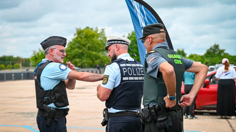 Un gendarme français, de profil à gauche, s'adresse à un policier allemand et un garde civil espagnol, vus de trois-quarts dos. Les trois hommes sont sur un parking, dont on distingue le bitume, avec une voiture rouge et deux personnes de dos à droite au second plan.