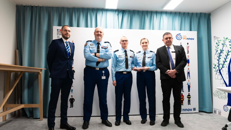 Trois officiers de gendarmerie, dont une femme tenant un troph&eacute;e, encadr&eacute;s par deux hommes en costume, posent devant un fond blanc sigl&eacute; lab MINNOV &agrave; droite.