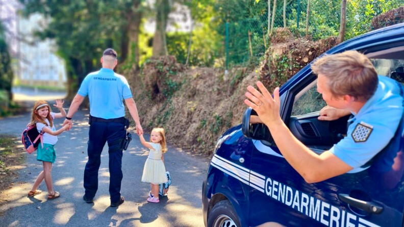 Agrandir l'image A doite, sortant de la fenêtre conducteur d'un véchiule de gendarmerie, un gendarme fait signe de la main à un autre gendarme de dos qui tient par la main deux fillettes avec un cartable
