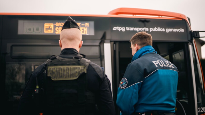 Un gendarme français et un policier suisse se tienennt debout, de dos, au niveau de la porte avant d'un autobus orange sur lequel est inscrite la mention " TPG - Transports publics genevois"
