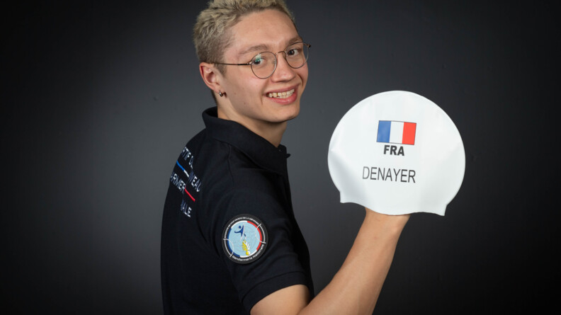 Agrandir l'image Un jeune homme blond à lunette, portant un polo noir et arborant l'écusson rond des sportifs de haut niveau de la Défense-gendarmerie, montre de sa main droite son bonnet de piscine siglé FRA avec un drpeau bleu-blanc-rouge et de son nom, Denayer