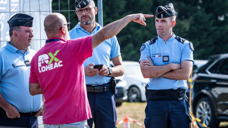 Trois gendarmes de face, regardent un homme de dos devant eux, au centre de l'image, vêtu d'un polo rose siglé "lohéac", montrant quelque chose au loin de son bras droit