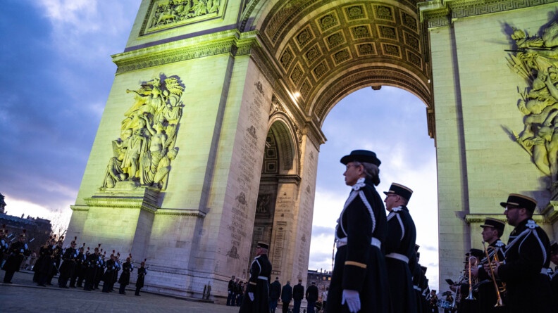 Vue en contre-plong&eacute;e de trois-quart sur l'Arc de Triomphe &eacute;clair&eacute;, tandis que la nuit tombe. Au premier rang, une rang&eacute;e de gendarmes au garde-&agrave;-vous. Derri&egrave;re eux des musiciens (trompettistes). Au second plan, les troupes au garde-&agrave;-vous.