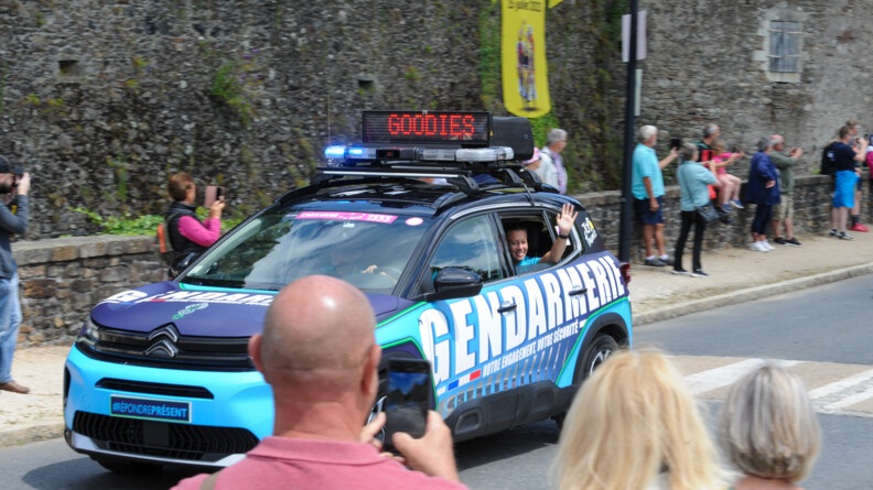 Agrandir l'image Un véhicule de la caravane gendarmerie du Tour de France avec les spectateurs qui filment sur les bas-côté. A la fenêtre arrière gauche du véhicule, une jeune gendarme souriante salue de la main