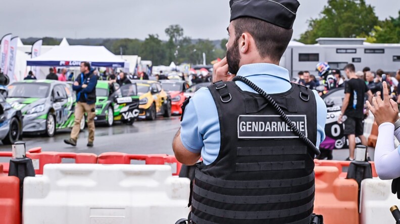 Un gendarme de dos derrière des barrières en plastiques rouge et blanche,limitant l'accès aux voitures de courses de diverses couleurs et ay public et coureurs sur place