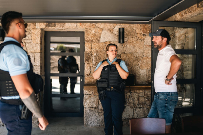 Agrandir l'image Deux gendarmes discutent avec un homme vêtu d'un jean, d'un t-shirt blanc et d'une casquette noire, se tenant à l'entrée d'une habitation