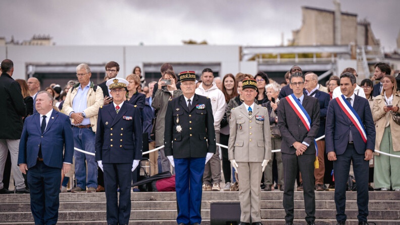 Des militaires hauts gradés, dont le directeur général de la gendarmerie nationale, se tiennent debout, aux côtés d'élus portant sur la poitrine l'écharpe tricolore. Derrière eux est installé un cordon de sécurité de couleur blanche, derrière lequel se trouve une foule.