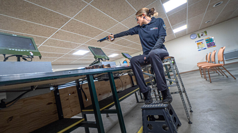 Dans un centre de tir en intérieur, Gaëlle Edon, sportive de la gendarmerie en para-tir, s'entraine. Elle est assise sur une sorte de chaise haute et tient son arme d'une seule main. Sur sa tenue sombre, on peut lire "sportive de haut niveau de la gendarmerie".