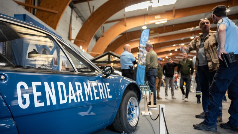 Vue sur le côté droit d'une ancienne ALpine de la gendarmerie, portant en blanc la mention "gendarmerie". A droite, et au fond, plus flous, des gendarmes échnagent avec des visiteurs
