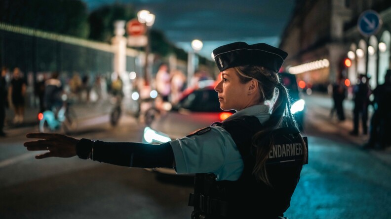 Une jeune femme gendarme se tenant debout, dans une rue de Paris, guide les voitures. Elle est équipée d'un gilet pare-balles portant dans le dos l'inscription "Gendarmerie".La scène se déroule la nuit.