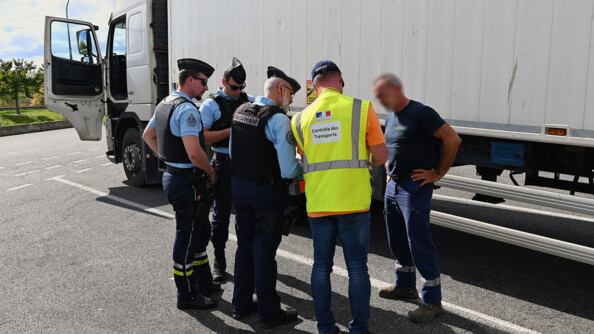 Trois gendarmes et un civil portant une chasuble jaune fluo "contrôle des transports" contrôlent un homme au pied d'un camion