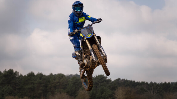 un motard de la gendarmerie vu de trois quart avant gauche et vu en conter-plongé suate dans les airs. En arrière-plan en bas, une forêt de résineux, en haut le ciel gris