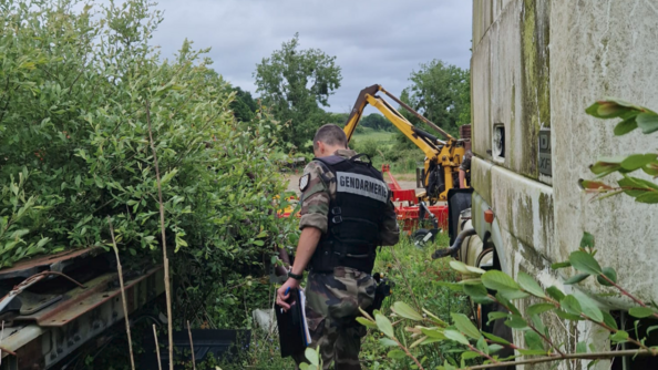 Un gendarme inspecte un terrain, au milieu d'une végération fournie. A sa droite se trouve le mur en béton d'un bâtiment, et plus loin, un engin agricole