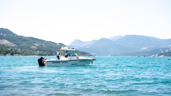 Une navette blanche portant l'insription GENDARMIE NATIONALE en lettres foncées, navigue sur un lac. Deux personnes se tiennent debout à son bord. Le lac est encerclé de reliefs.