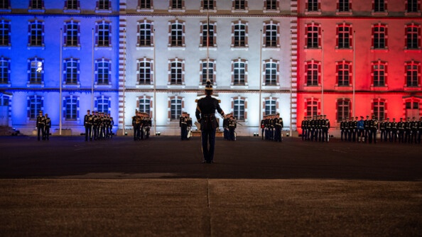De dos un cadre de l'EOGN salue les officiers issus du rang alignés devant l'état-major de l'Ecole illuminée de bleu blanc rouge