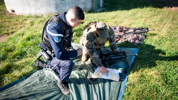 Un gendarme et un militaire de l'armée de terre, tous deux en tenue, accroupis côte à côte sur une bâche disposée dans l'herbe, regardent une carte. Le militaire de l'armée de terre désigne du doigt un point sur la carte. Devant lui est posé son fusil.
