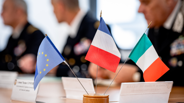 Gros plan sur trois petits drapeaux, européen, français et italien, posés sur une table de réunion, où se trouvent également des cartouches portant le nom des participants. En arrière plan, flous, trois officiers en vareuse.