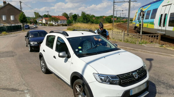 Un gendarme dispense des conseils de pr&eacute;vention au conducteur d'un v&eacute;hicule blanc arr&ecirc;t&eacute; &agrave; un passage &agrave; niveau, tandis qu'un train passe. Au second plan, un second v&eacute;hicule noir.