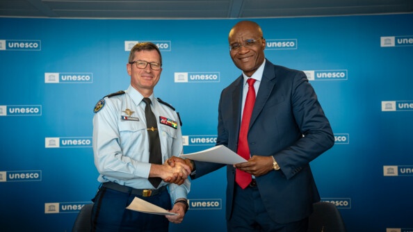 A gauche un général de gendarmerie et à droite un homme en civil qui tient un document, se serrent la main en souriant. Au fond, un mur bleu portant à de nombreuses reprise le logo de l'UNESCO