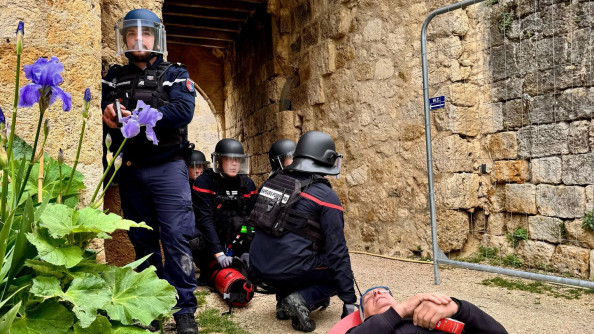 Un gendarme casqué à un angle de rue, debout, derrière lui et à sa gauche, quatre polmpiers un genoux au sol, casqué, devant eu une victime fictive sur un brancard rouge, sous une rue