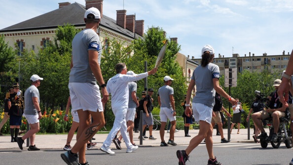 Un homme vétu de blanc élève la flame olympique, de ârt et d'autre trois hommes vêtus d'un polo bleu et short beige. En fond la grille de l'Ecole des officiers de la gendarmerie nationale