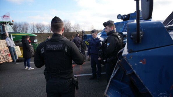 Trois gendarmes devant des véhicules blindés, regardent les agriclteurs et leur tracteur qui leur font face.