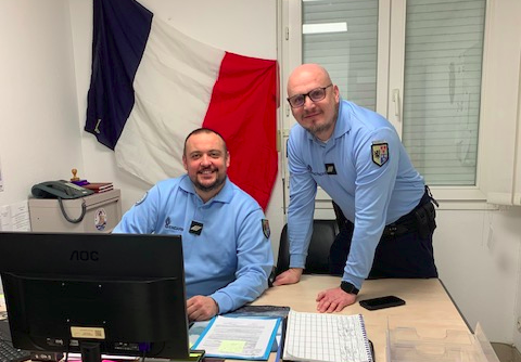 Deux gendarmes souriants se tiennent derrière un bureau. L'un 'eux est assis, devant un écan d'ordinateur, tandis que le second se tient de bout à ses côtés, les mains posées sur le bureau, le corps légèrement incliné en direction de son collègue. Derrière eux, sur le mur blanc, est érigé un grand drapeau français.