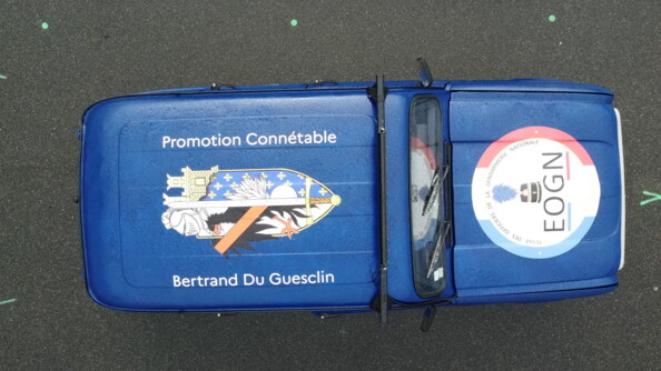 On aper&ccedil;oit vue d'en haut une Renaud 4L bleu marine, couverte d'une grande enseigne sur le toit avec la mention "Promotion Conn&eacute;table Bertrand Du Guesclin", et une seconde sur le capot portant la mention "Ecole des officiers de la gendarmerie nationale EOGN"