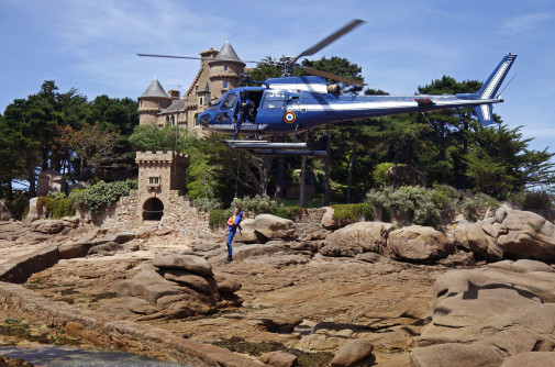 Un h&eacute;licopt&egrave;re bleu marine de la Gendarmerie nationale est en vol stationnaire au-dessus d&rsquo;un littoral rocheux. Un plongeur en tenue de sauvetage est suspendu sous l'appareil, au dessus des rochers. Le paysage est constitu&eacute; de gros rochers arrondis, et d'un petit ch&acirc;teau au milieu d'un bosquet d'arbres sous un ciel bleu l&eacute;g&egrave;rement nuageux.