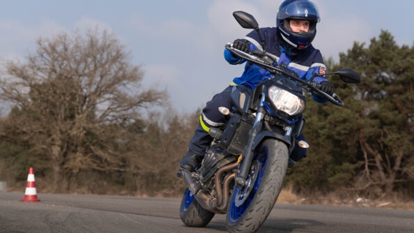 un motard de la gendarmerie sur sa moto, vue en contre-plongée de trois quart avant droit, porte le regard au loin. Il est sur un chemin goudronné. Au fond à gauche, un plot orange et blanc. EN arrière-plan, flou, des arbres