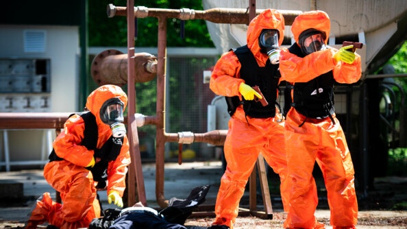 Un gendarme en tenue NRBC est agenouillé auprès d'un gendarme victime allongé sur le dos. Deux autres gendarmes en tenu NRBC ont en main leur pistolet et restent en appui.