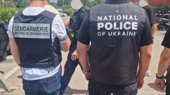 De dos à gauche, un enquêteur en civil portant au bras droit un brassard gendarmerie et une chasuble noire "gendarmerie section de recherches" et à droite, de dos, un enquêteur arborant une chasuble noire "national police of ukraine"