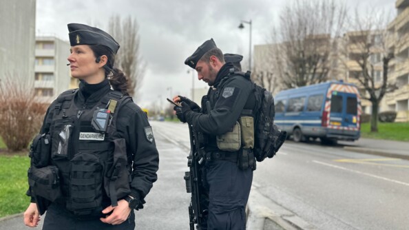 Une femme gendarme en tenue bleue marine avance dans la rue, face &agrave; l'objectif. Derri&egrave;re elle se trouvent deux autres gendarmes, dont un consultant un smartphone. Tous se trouvent au coeur d'un ensemble d'immeubles d'habitation. On aper&ccedil;oit &eacute;galement en arri&egrave;re plan un fourgon bleu de la gendarmerie, stationn&eacute; le long du trottoir.