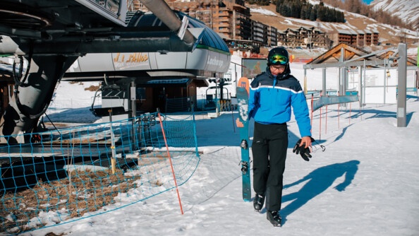une femme gendarme tenant deux skis de la main droite avance sur la neige, en fond une station de ski