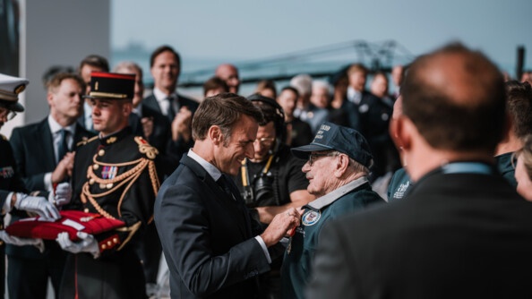 Le président de la République remet une médaille à un vétéran. Dans le fond, parmi la foule, on observe des gendarmes de la garde Républicaine qui tiennent les coussins rouges sur lesquels sont posées les médailles.
