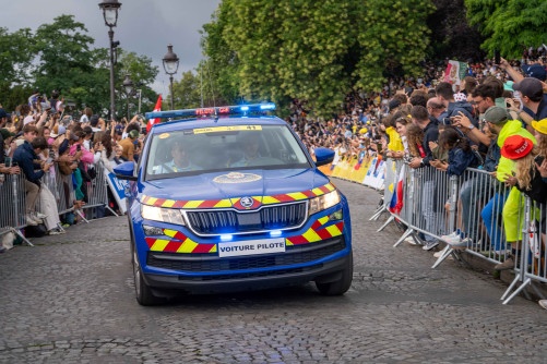 Une voiture de la gendarmerie roule dans les rues de Paris, sur une voie pavée, le logn de laquelle est massée une foule imortante, contenue derrière des barrières