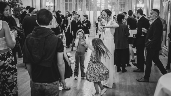 Scèe de fête avec des enfants dansants et adultes conversant, un verre à la main. Au milieu, une femme vêtue d'une combinaison blanche danse avec les enfants