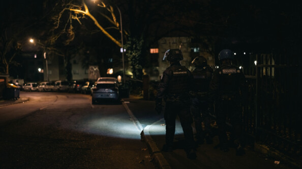 trois gendarmes casqués marchent dans la rue. Ils s'éclairent avec une lampe torche parce qu'il fait nuit et sombre.