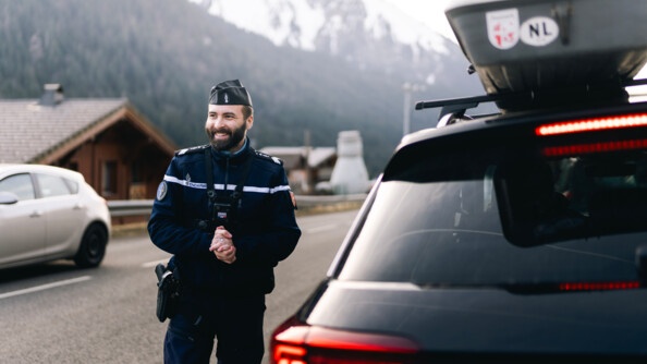 Image d'un gendarme souriant, debout près d'une voiture noire, sur le toit duquel est accrochée une galerie. Le militaire vient de procéder à un contrôle du véhicule et du conducteur.