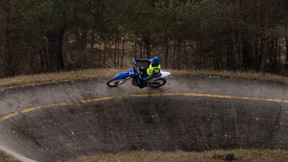Un motard de la gendarmerie sur une moto cross roule sur la partie haute d'un virage très serré en hauteur et goudronné. Il est marqué dun' ligne jaune. En arrière plan, une forêt de résineux