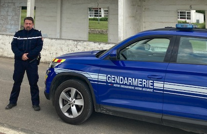 Un homme d'âge moyen, en uniforme de gendarme, pose debout, les mains croisées devant lui, à côté de son véhicule bleu. Sur la portière, la mention "Gendarmerie" est inscrite