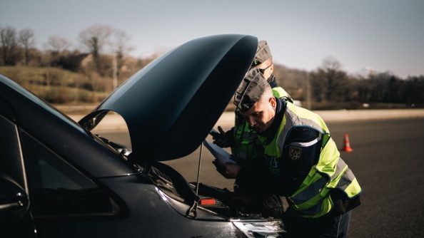 Deux gendarmes inspectent le moteur d'un véhicule noir, dont le capot est relevé.