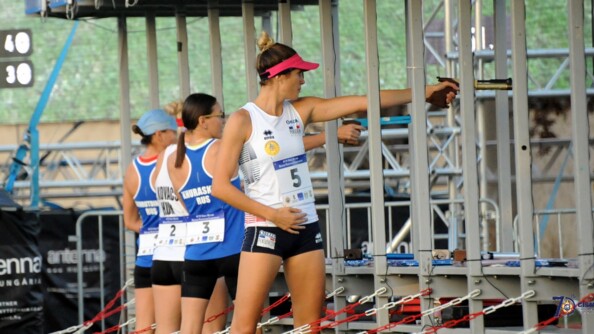 Quatre femmes de dos, la première de profil, en tenue de sport devant des box de tir en métal, visent pour du tir sportif