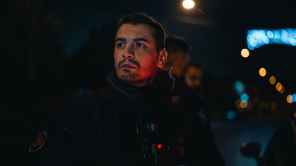 Dans la nuit, il fait sombre, un gendarme regarde droit devant lui. Derrière est en train d'être réalisé un controle routier.