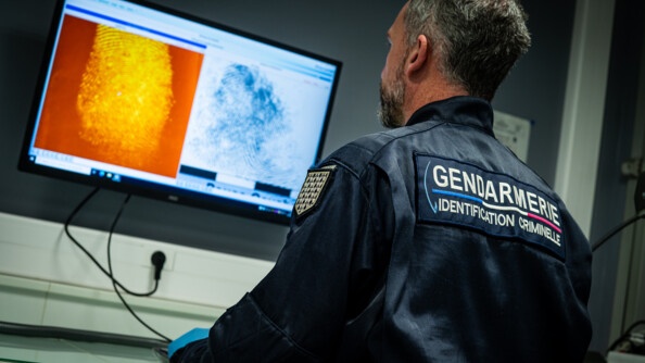 Un gendarme de dos, en bleu marine, portant la mention "gendarmerie, identification criminelle", observe un écran d'ordinateur accroché au mur, exposant une empreinte digitale orange et une bleue