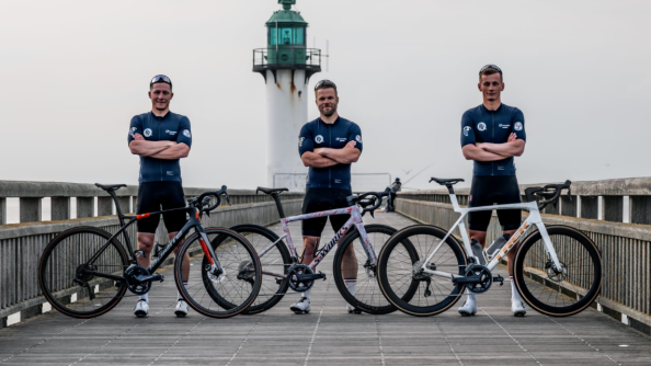 Trois hommes en tenue de sport (haut bleu marine et short noir), sur un pont au bot duquel se trouve un phare blanc et vert, debout derrière leurs vélos