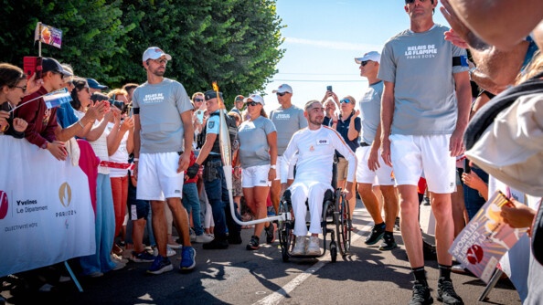 Personne à mobilité réduite porteuse de la flamme accrochée à son fauteuil dans les Yvelines, sous la protection de gendarmes placés le long du parcours.